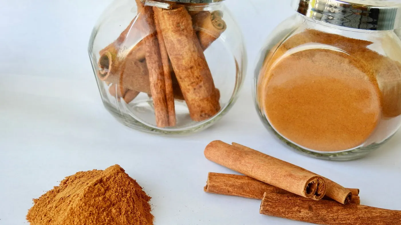 Tres tubos de canela en rama y un montoncito de canela molida junto a dos frascos de vidrio con canela, sobre una mesa blanca