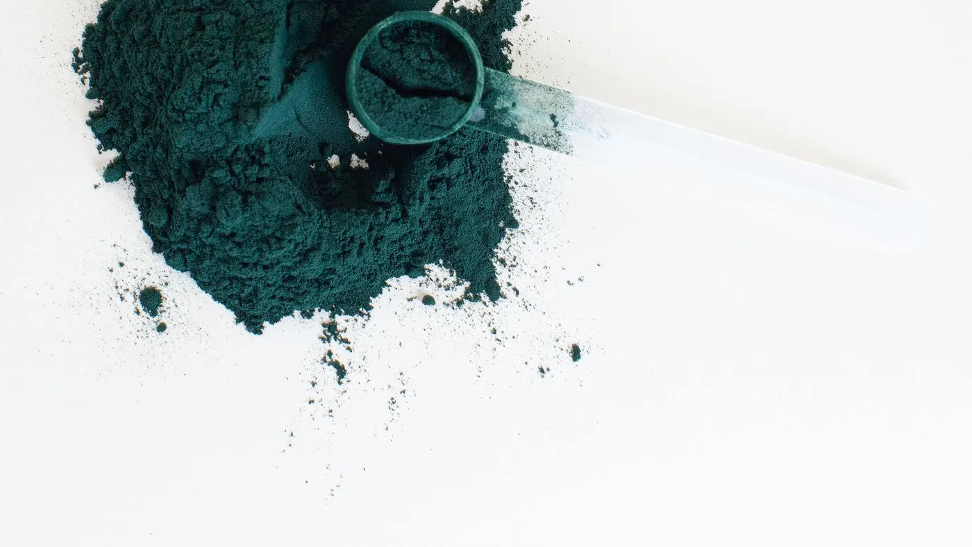 Polvo verde oscuro de espirulina visto desde arriba, con una cucharita medidora transparente sobre fondo blanco